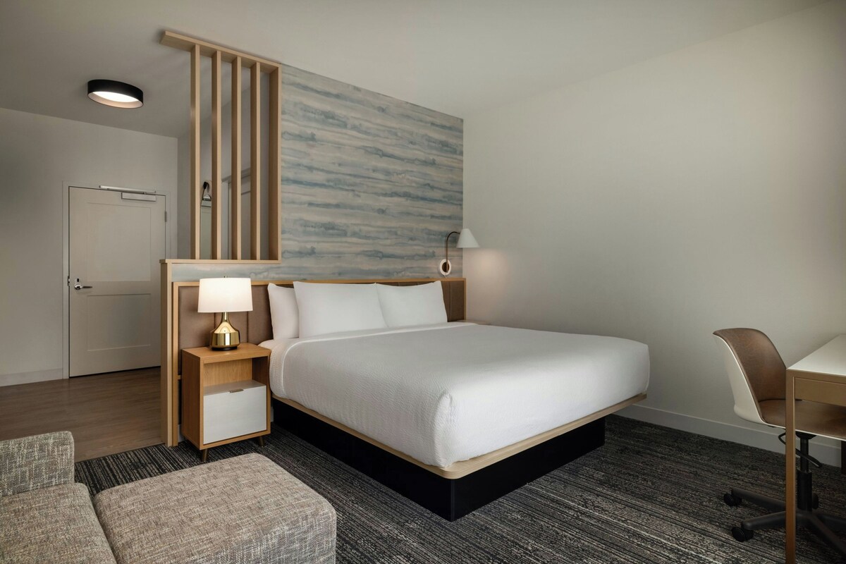 A hotel room with a bed, bedside tables, a desk, a chair, and a large abstract wall painting.