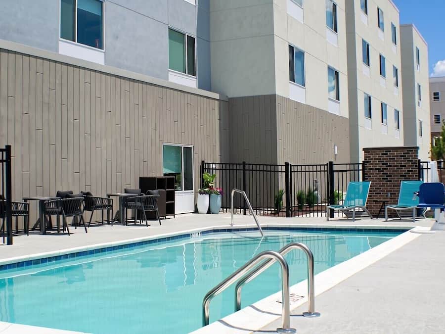 Piscina com escada de metal, rodeada por espreguiçadeiras e mesas, com um edifício de design moderno ao fundo.