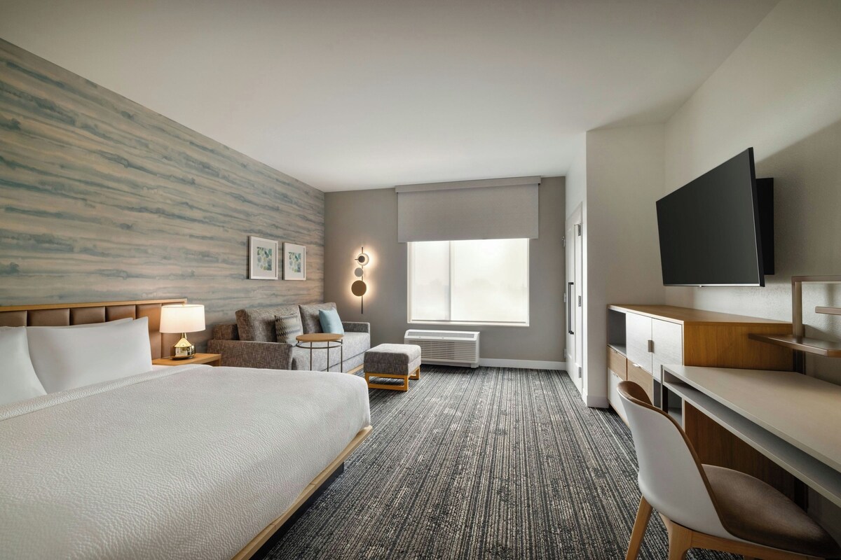 A hotel room with a large bed, a desk, a chair, a TV, a sofa, a small table, a lamp, a wall-mounted light, and a window with a view.