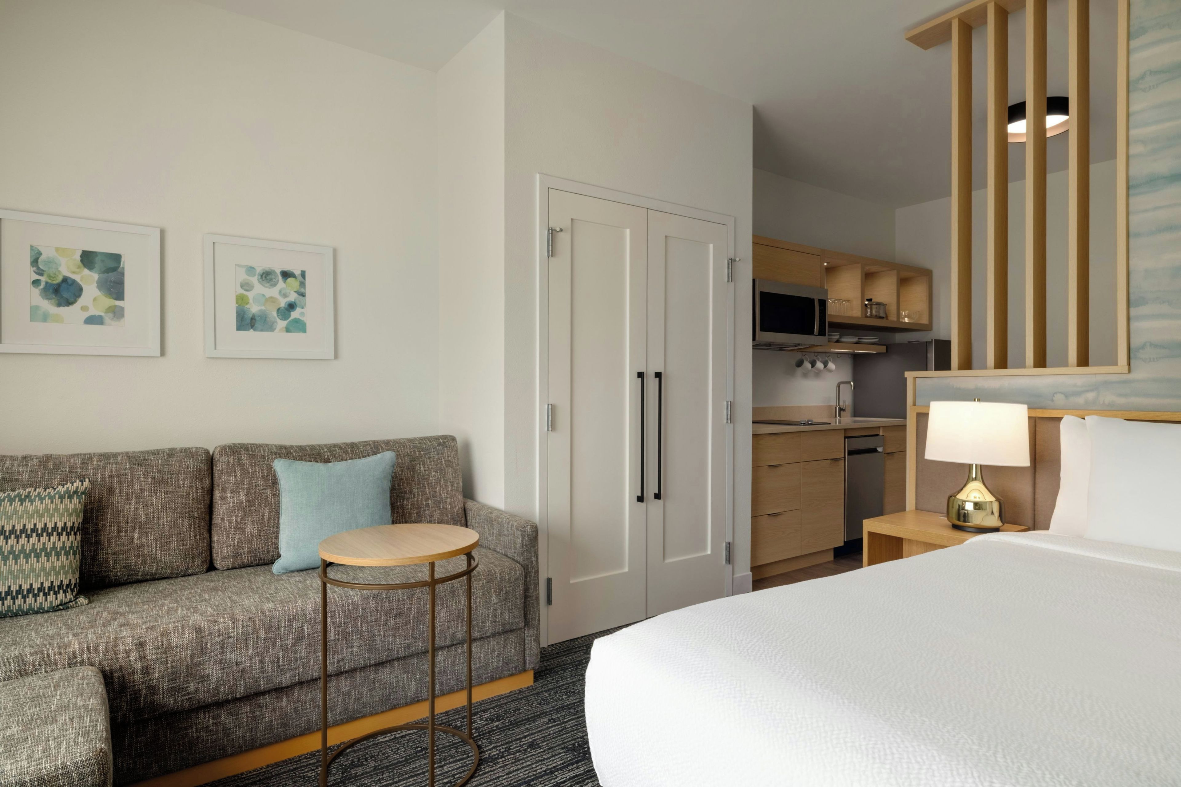 A hotel room with a bed, a sofa, a small round table, a kitchenette, and artwork on the wall.