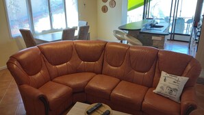 A brown leather sectional sofa with a remote control on a small wooden table, a dining area with chairs, and a kitchen visible through an open door. - Between river and town center - air-conditioned - 2 WCs - enclosed courtyard (Cognac)