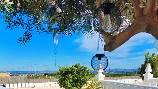 Un'area salotto esterna con sedute bianche, cuscini viola, una sedia in vimini e una pianta in vaso.