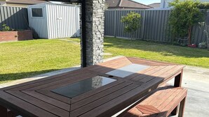A wooden picnic table with a glass top, set on a concrete patio with a stone pillar, surrounded by a backyard with a wooden shed and a fence. - Modern Napier Comfort (Napier)