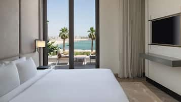 Habitación de hotel con una cama grande, un televisor montado en la pared, vista a la playa y un balcón con mesa y sillas.