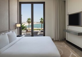Habitación de hotel con una cama grande, un televisor montado en la pared, vista a la playa y un balcón con mesa y sillas.