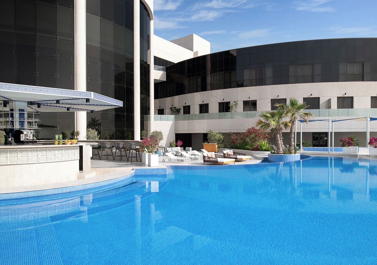 A swimming pool with lounge chairs and a bar area, surrounded by a modern building with large windows.