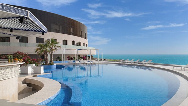 A swimming pool with a curved design, a building with a modern facade, and a view of the ocean. - Ray Hotel by Cloud 7 (Mangaf)
