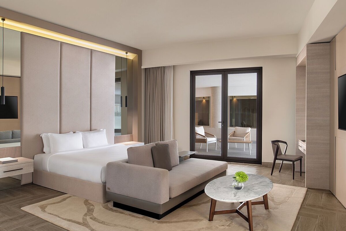 Une chambre d’hôtel moderne, équipée d’un lit, d’un canapé, d’une table ronde et offrant une vue à travers une porte vitrée.
