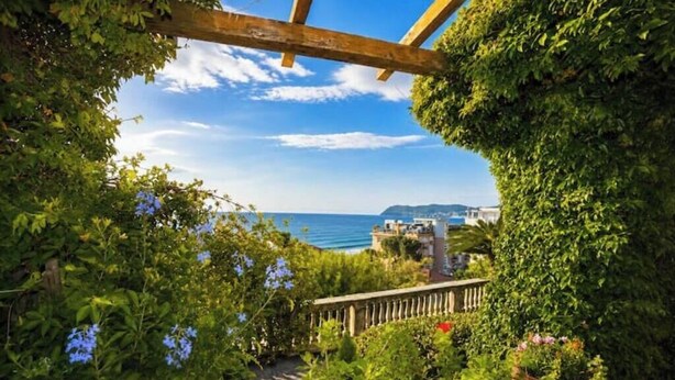 A garden with a wooden pergola, lush greenery, and a view of the sea and buildings. - Hostdomus - Villa Gabriella (Alassio)
