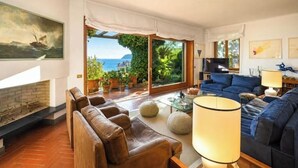 A living room with a fireplace, a blue sofa, a coffee table, and a view of the sea through large windows. - Hostdomus - Villa Gabriella (Alassio)