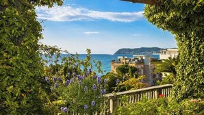 A coastal view with a wooden pergola, lush greenery, and a building with balconies overlooking the sea. - Hostdomus - Villa Gabriella (Alassio)
