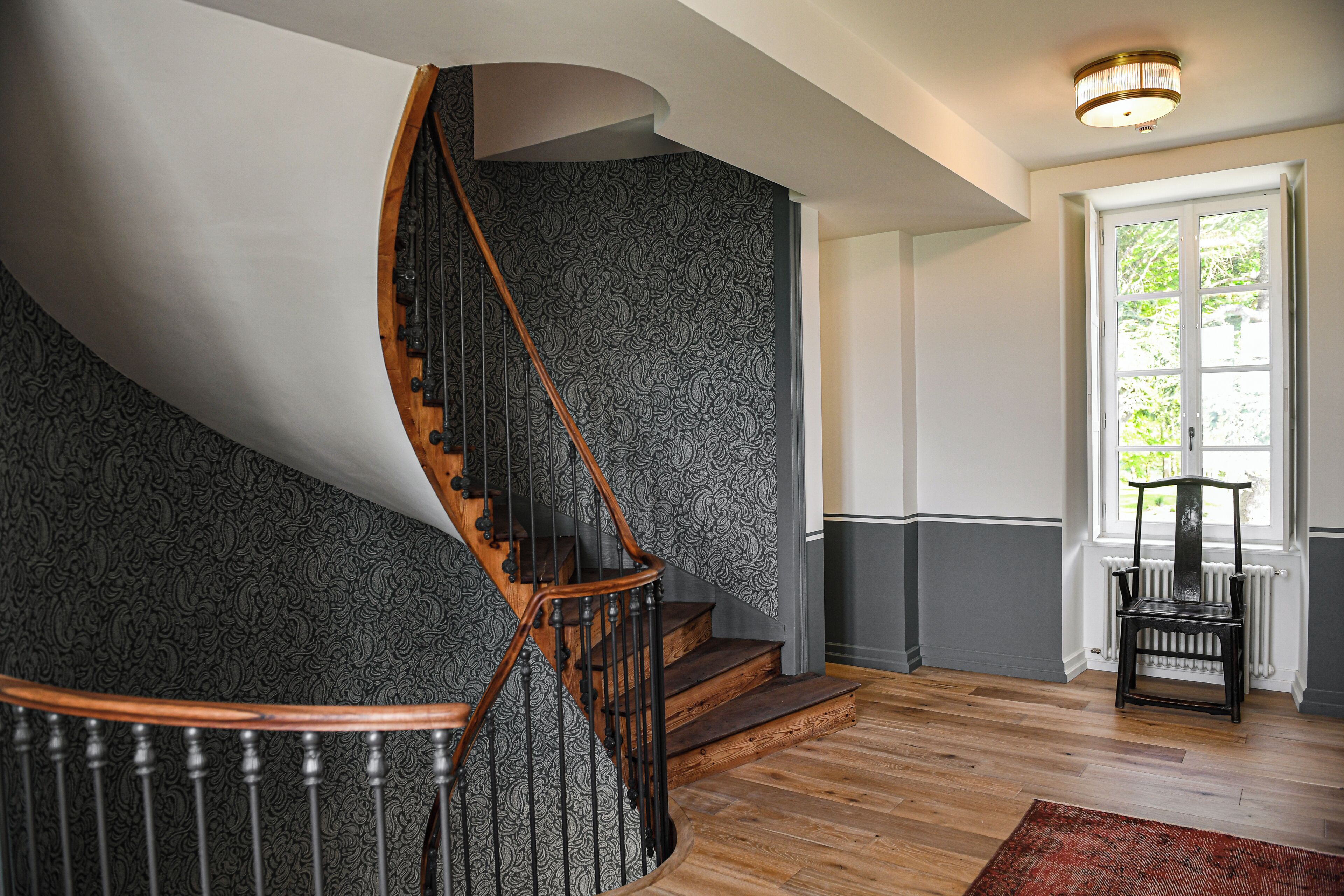 A spiral staircase with a wooden handrail and black metal balusters, leading to a landing with a view of a window and a radiator.