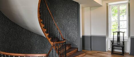 A spiral staircase with a wooden handrail and black metal balusters, leading to a landing with a view of a window and a radiator.