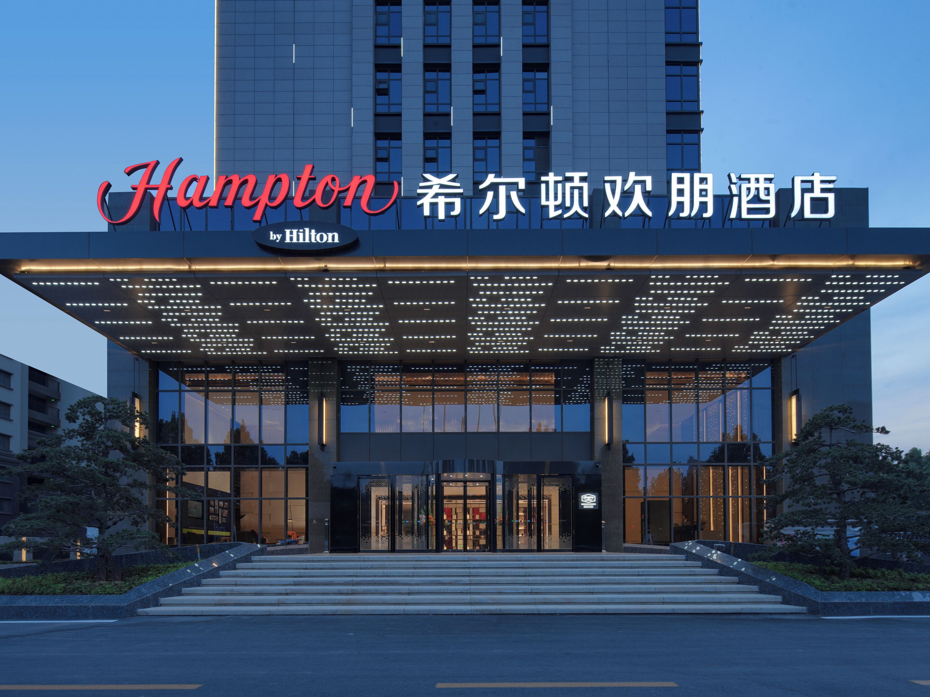 A hotel entrance with a modern design, featuring a large illuminated sign, a covered entrance area, and a staircase leading to the lobby.