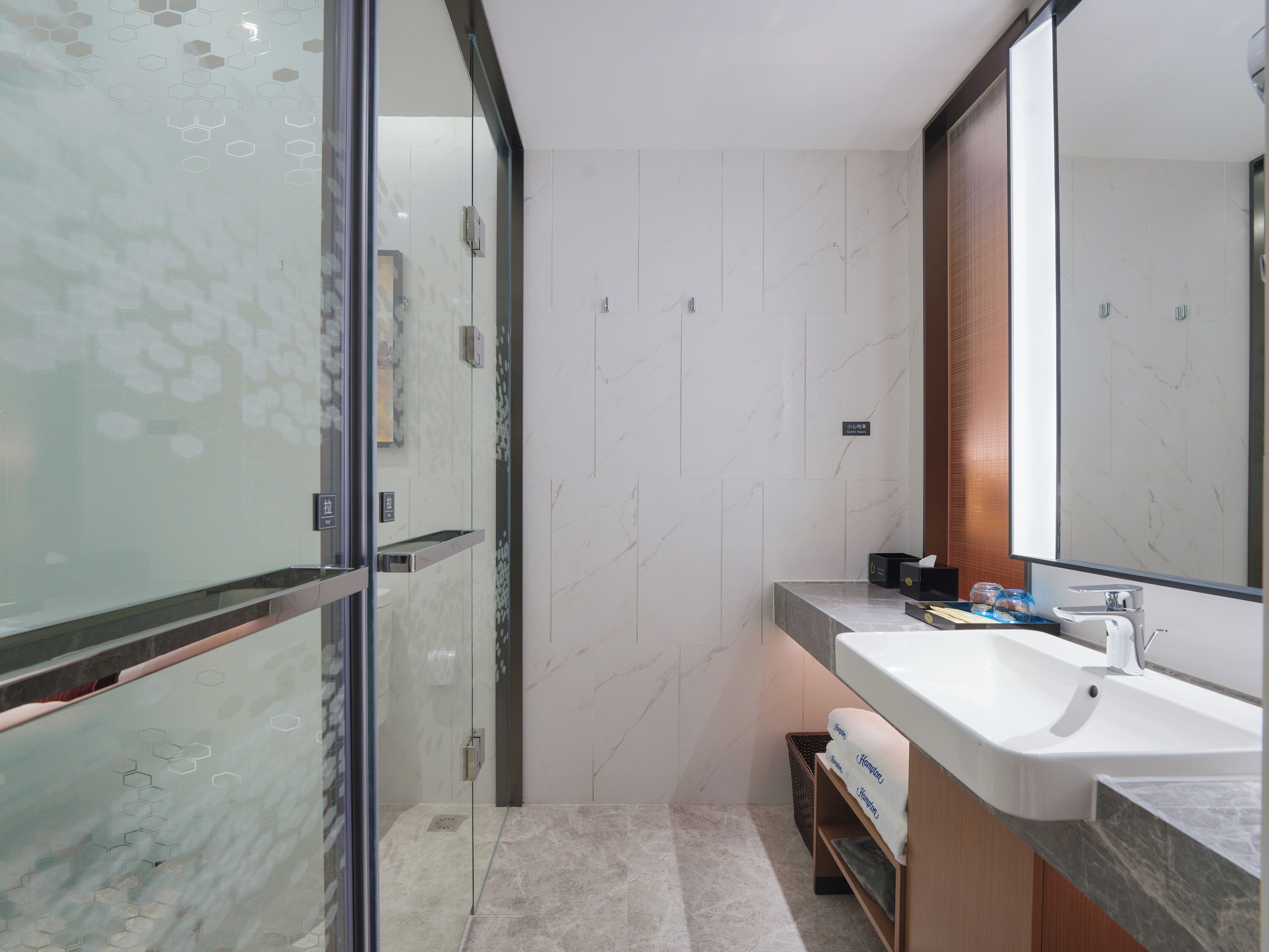 A modern bathroom with a glass shower enclosure, a marble-tiled wall, a large mirror, and a sink with a towel rack.