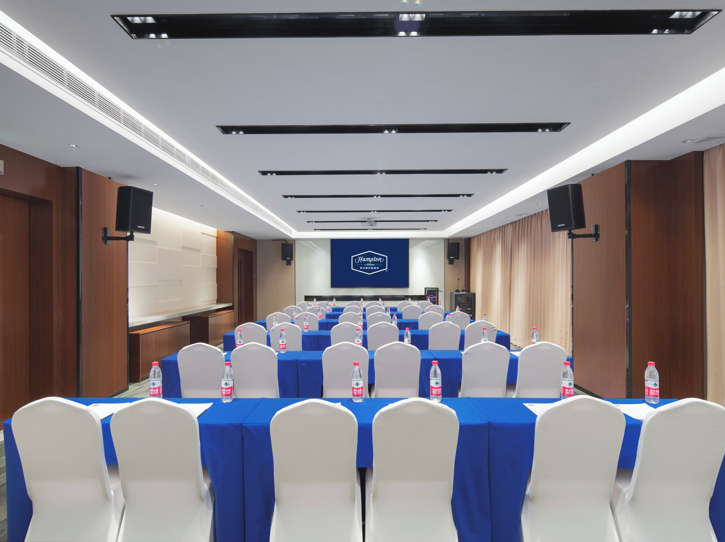 Une salle de conférence avec des rangées de tables, des chaises blanches, des nappes bleues et des bouteilles d’eau.