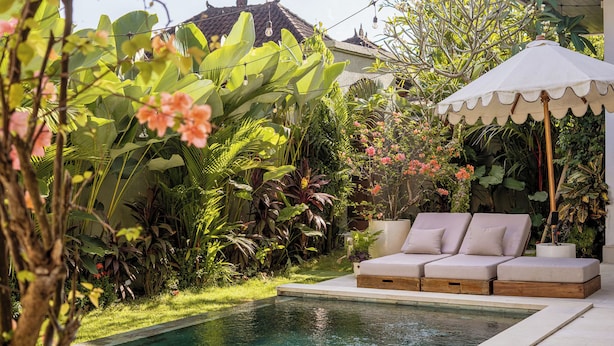 Zona junto a la piscina con una silla de descanso y sombrilla, rodeada de exuberante vegetación y una piscina.