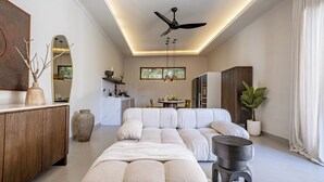 Una sala de estar moderna con un sofá seccional, ventilador de techo, luces colgantes y un amplio ventanal.