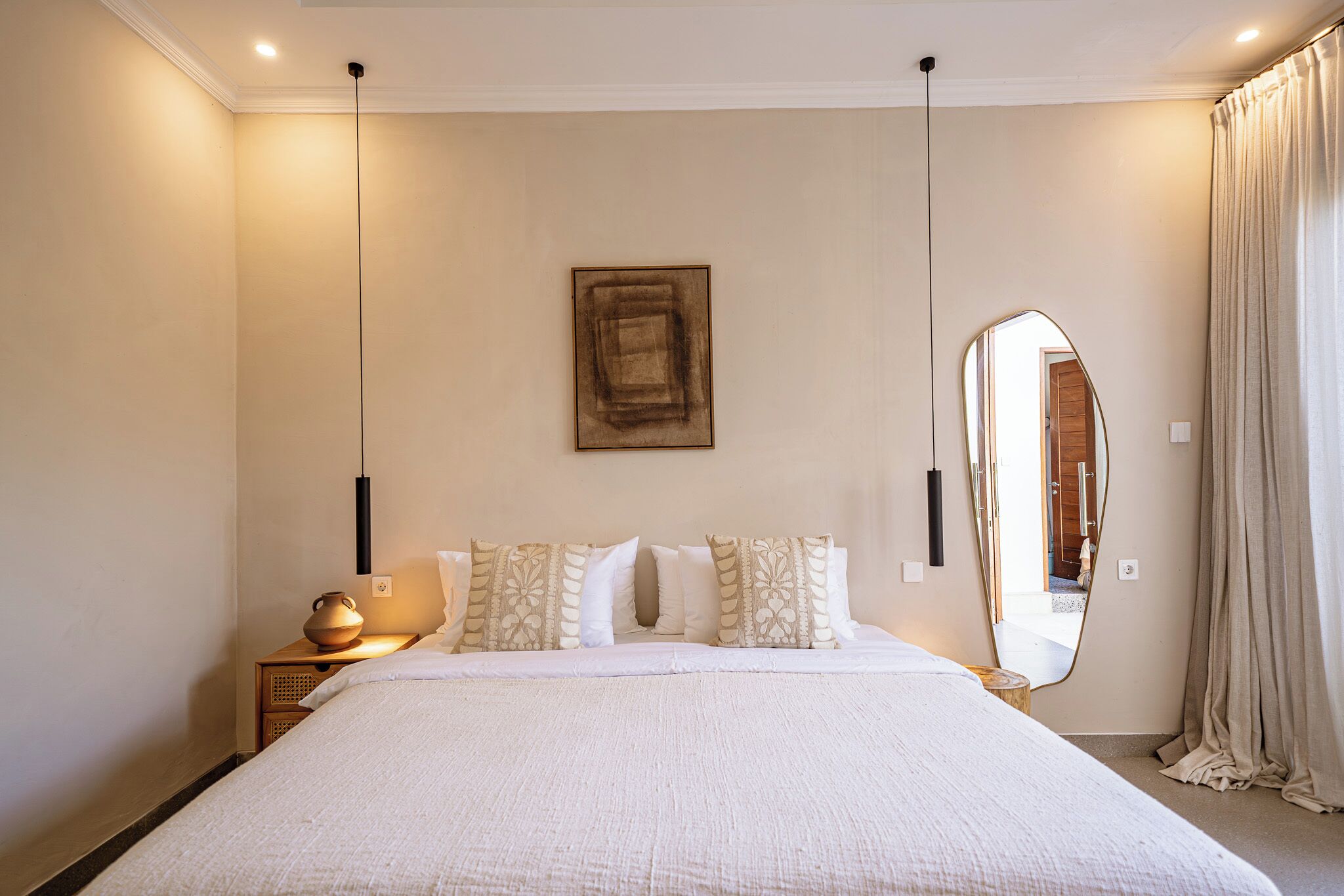 Une chambre à coucher avec un grand lit, une table de chevet avec une lampe, un miroir et une œuvre d’art encadrée accrochée au mur.