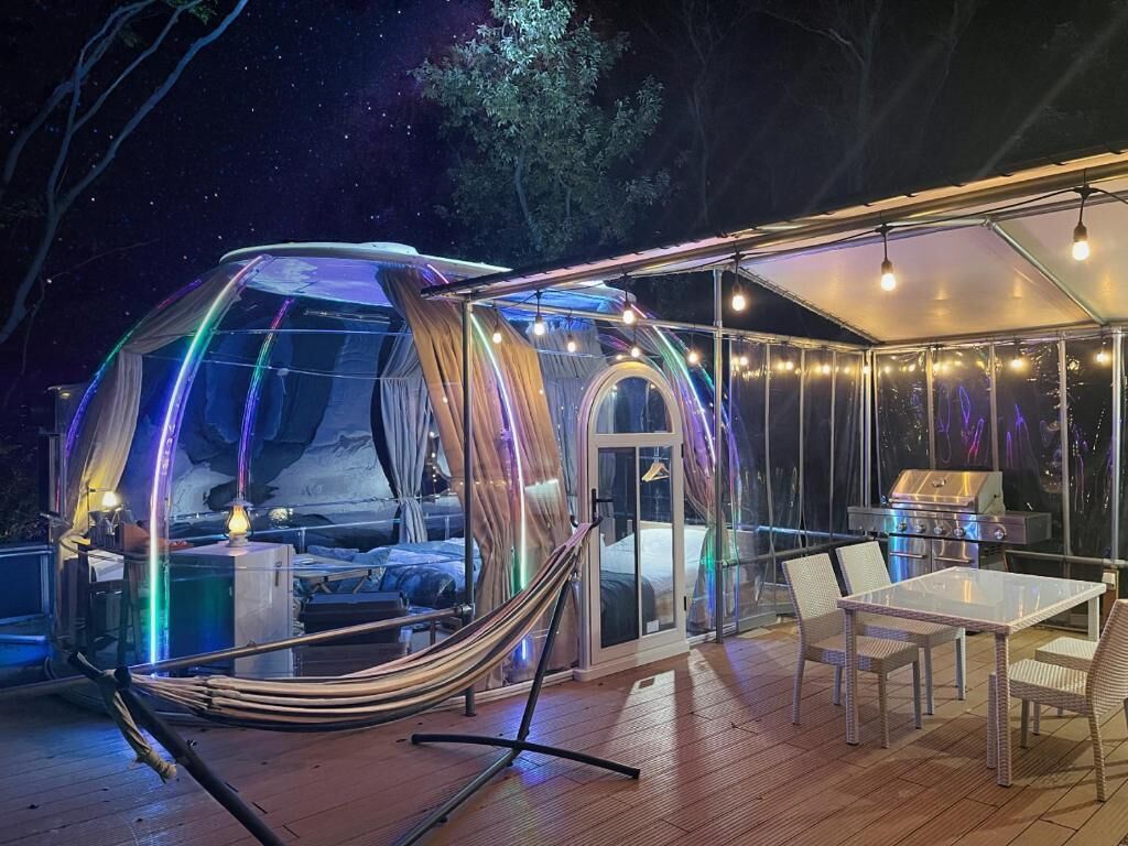 A geodesic dome structure with a transparent floor, a hammock, a table, and chairs.