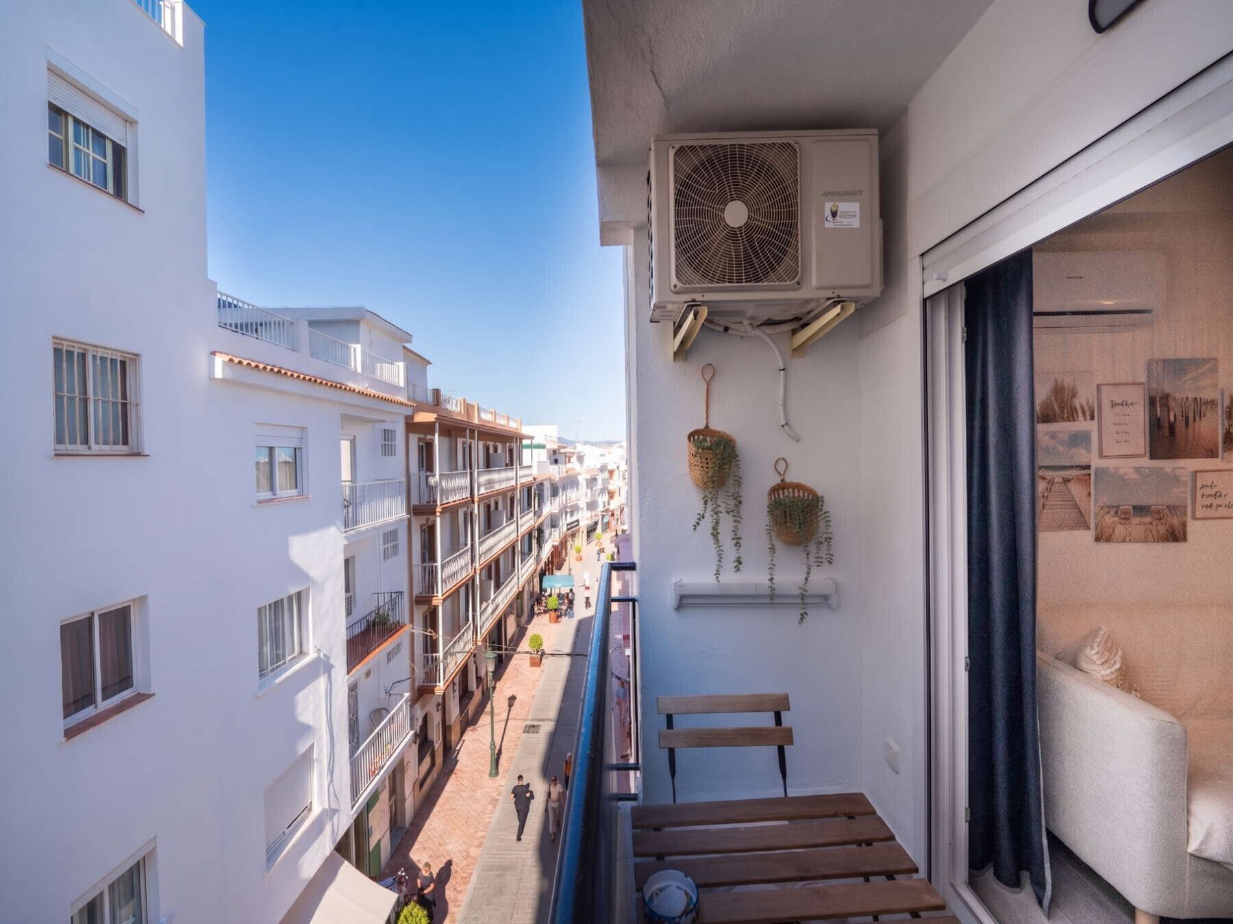 Un balcone con vista sulla strada, un condizionatore d'aria e cesti fioriti appesi.