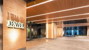 Entrada moderna de um edifício com uma placa indicando 'JEWEL OKLEY KUALA LUMPUR', apresentando um piso de pedra polida, teto de painéis de madeira e portas de vidro que dão acesso ao interior.