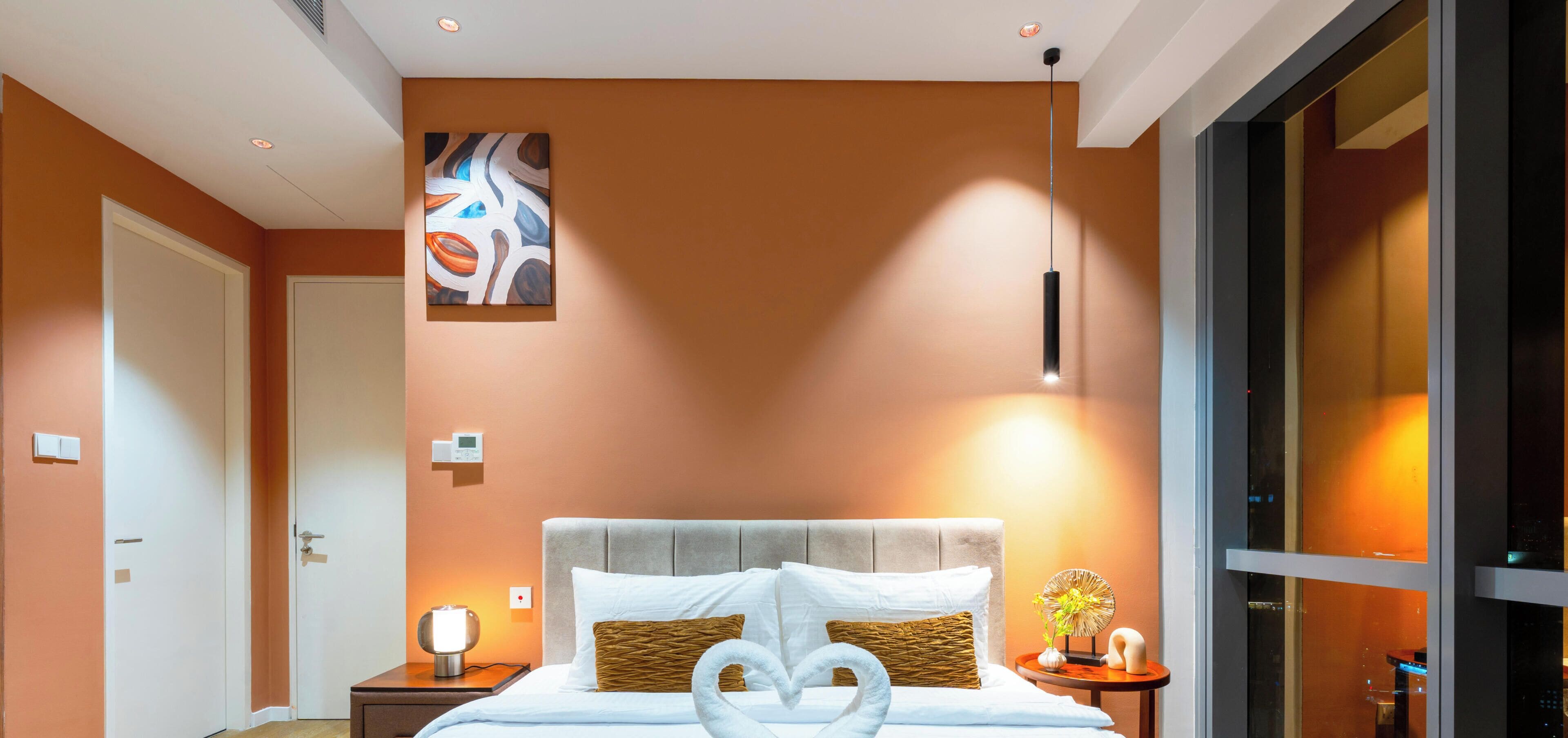A modern hotel room with a large bed, a bedside table with a lamp, a wall-mounted artwork, and a large window.