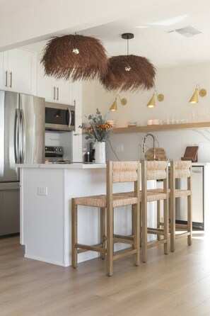 A modern kitchen with a central island, wooden chairs, and pendant lights. - The SouthOhana Beach House with Ocean Views! (Oceanside)