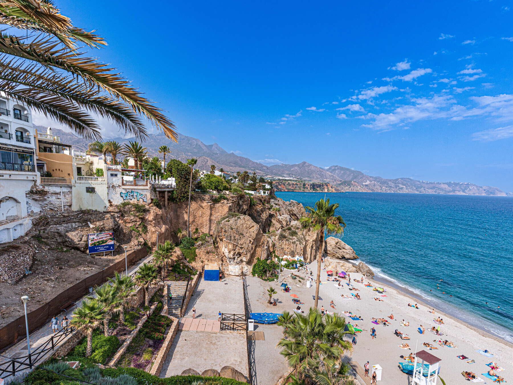 A coastal beach with a rocky cliff, palm trees, and a clear blue sea.