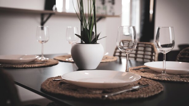 Une table à manger dressée avec des assiettes, des couverts et des verres à vin, et un vase de fleurs placé au centre.