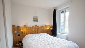 Une chambre à coucher avec une tête de lit en bois, un couvre-lit blanc, une table de chevet avec une lampe et une fenêtre donnant sur des immeubles.