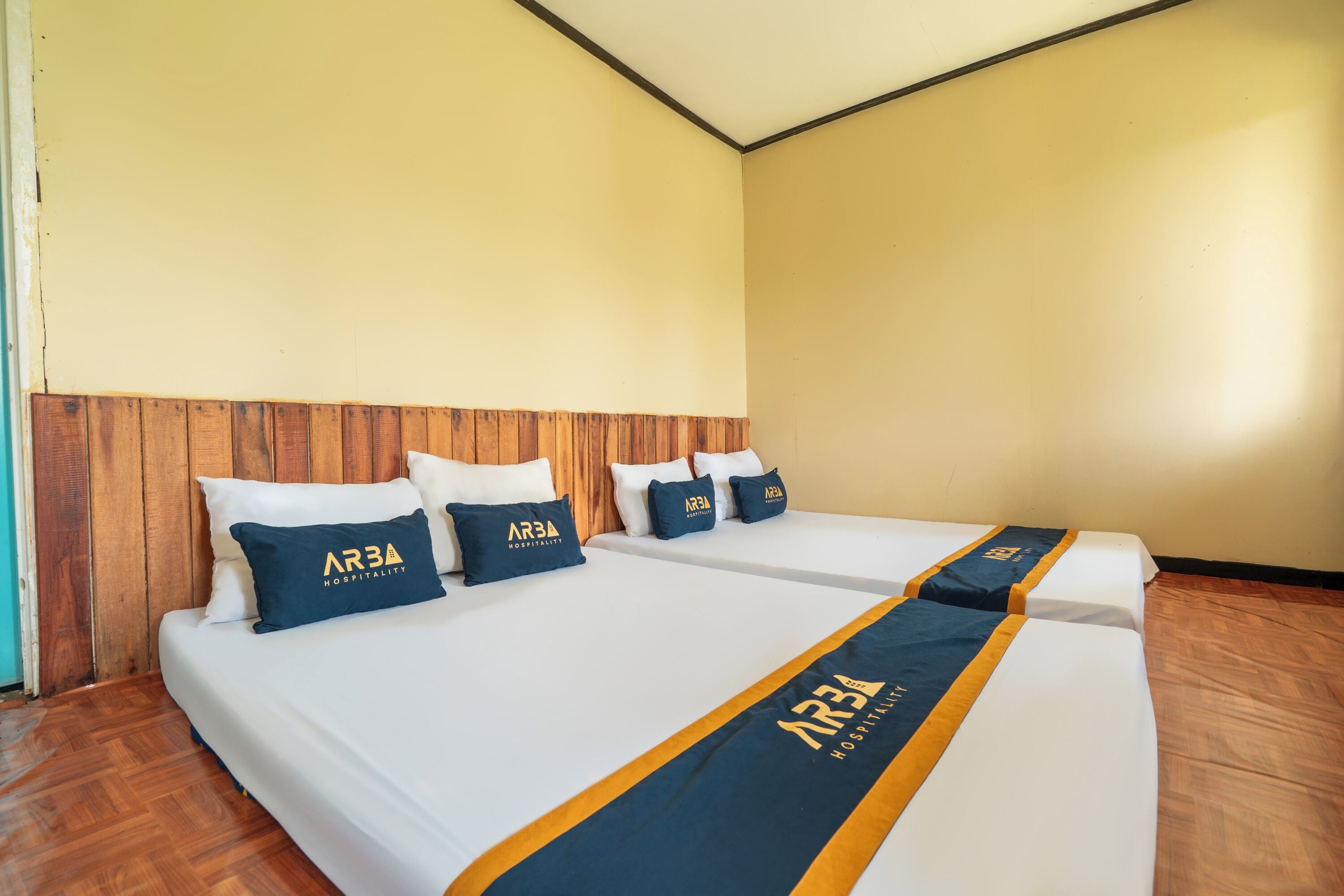 Habitación de hotel con dos camas, cabecero de madera, ropa blanca y almohadas azules con el logotipo de 'AR3A Hospitality'.