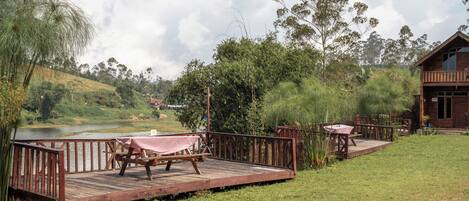 A wooden deck with a picnic table and chairs overlooking a lake, surrounded by lush greenery and a wooden cabin.