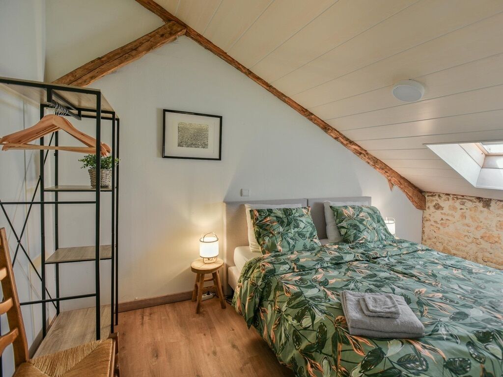 Ett sovrum med ett sluttande tak, en säng med ett sängöverkast med ett lövmönster, en trästol, ett litet nattduksbord med en lampa och en inramad tavla på väggen.