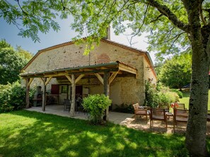 Ein Steingebäude mit einer überdachten Terrasse, Sitzgelegenheiten im Freien und einem Garten.