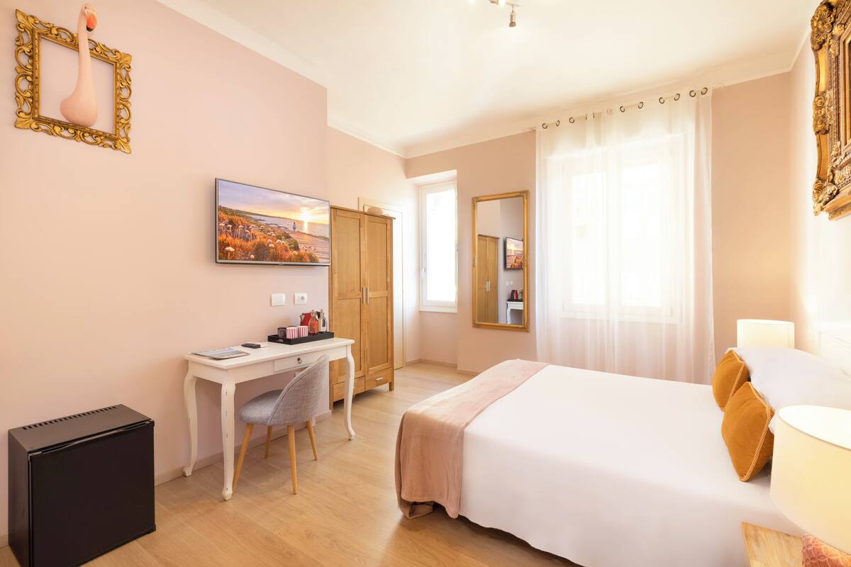 A hotel room with a bed, a desk, a chair, a wardrobe, a mirror, and a painting on the wall.
