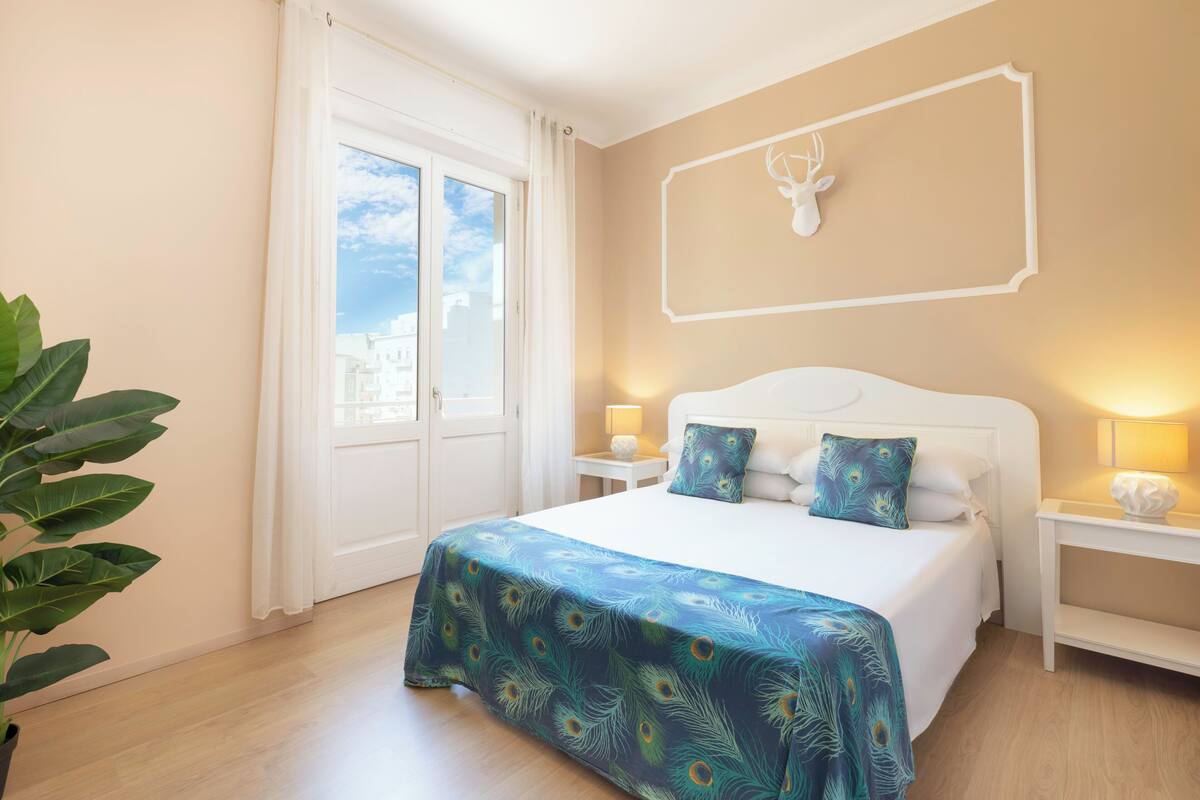 A bedroom with a large bed, a nightstand with a lamp, a wall-mounted deer head decoration, and a view of buildings through a door.