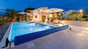 En swimmingpool med liggestole, et overdækket siddeområde og et moderne hus i baggrunden.