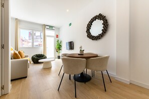 Ein modernes Hotelzimmer mit einem runden Esstisch, zwei Stühlen, einer Couch mit Kissen, einem grünen Sitzsack, einer Topfpflanze, einem an der Wand montierten Fernseher und einem dekorativen Kranz.