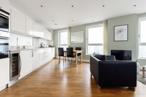 Eine moderne Küche mit weißen Schränken, ein Essbereich mit Tisch und Stühlen, ein schwarzes Sofa und ein Fernseher in einem Wohnbereich mit großen Fenstern.