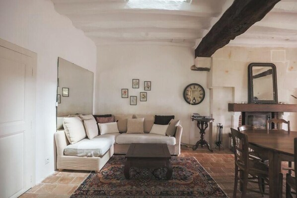A living room with a sofa, coffee table, and a dining table with chairs. There is a clock on the wall and a fireplace.