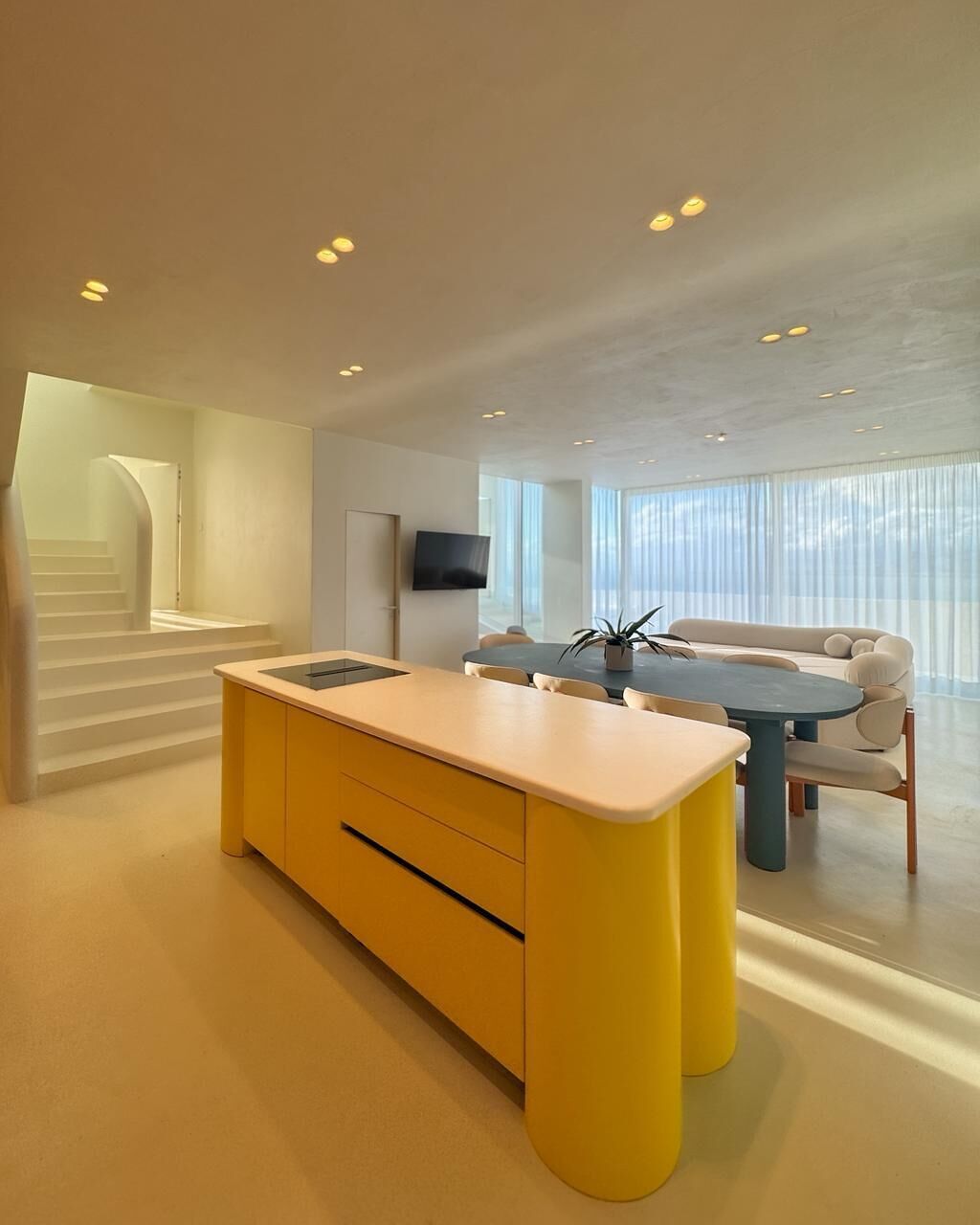 A modern kitchen with a central island, a dining area with a round table, and a staircase leading to an upper level.
