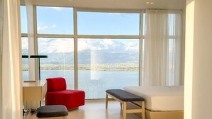 Una habitación de hotel moderna con un ventanal grande, una silla roja, un banco de madera y una cama.