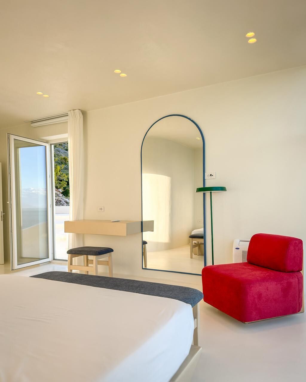 A modern hotel room with a bed, a red chair, a large mirror, and a view through a glass door.