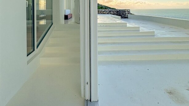 Un edificio moderno con fachada de vidrio, un balcón con vistas al mar y un muro de piedra al fondo.
