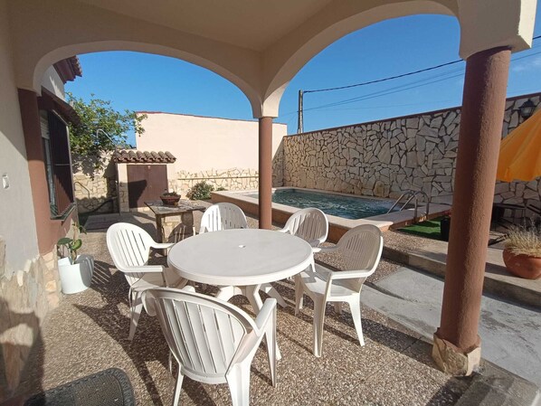 An outdoor patio with a round table and white chairs, a stone wall, and a swimming pool in the background.