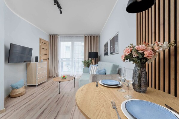 A modern living room with a flat-screen TV, a dining table set with plates and cutlery, a sofa, a coffee table, and a vase with flowers.