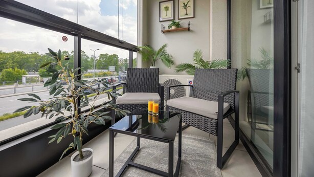 A balcony with a glass table, two wicker chairs, potted plants, and framed pictures on the wall. - Bright Apartment with Parking and Balcony | Close to the Airport (Warszawa)
