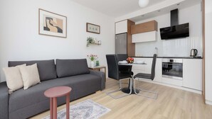 A modern living room with a grey sofa, a small round table, and a kitchen area with white cabinets and a black range hood. - Bright Apartment with Parking and Balcony | Close to the Airport (Warszawa)