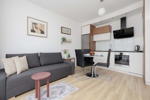 A modern living room with a grey sofa, a small round table, and a kitchen area with white cabinets and a black range hood. - Bright Apartment with Parking & Balcony Close to the Airport (Warszawa)